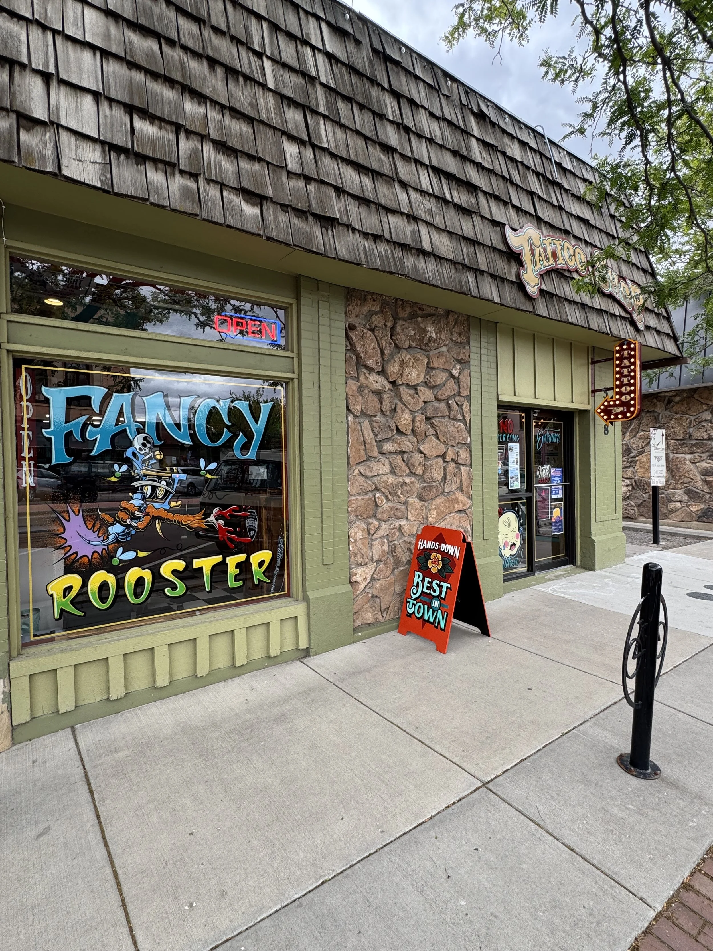 View of a storefront with a wood shingle roof, display window with cartoon skeleton holding a guitar, neon 'Open' sign, a decorative sign that reads 'Tattoo', and a sandwich board sign that says 'Hands Down, Best in Town'. Sidewalk and some green trees are in the foreground.