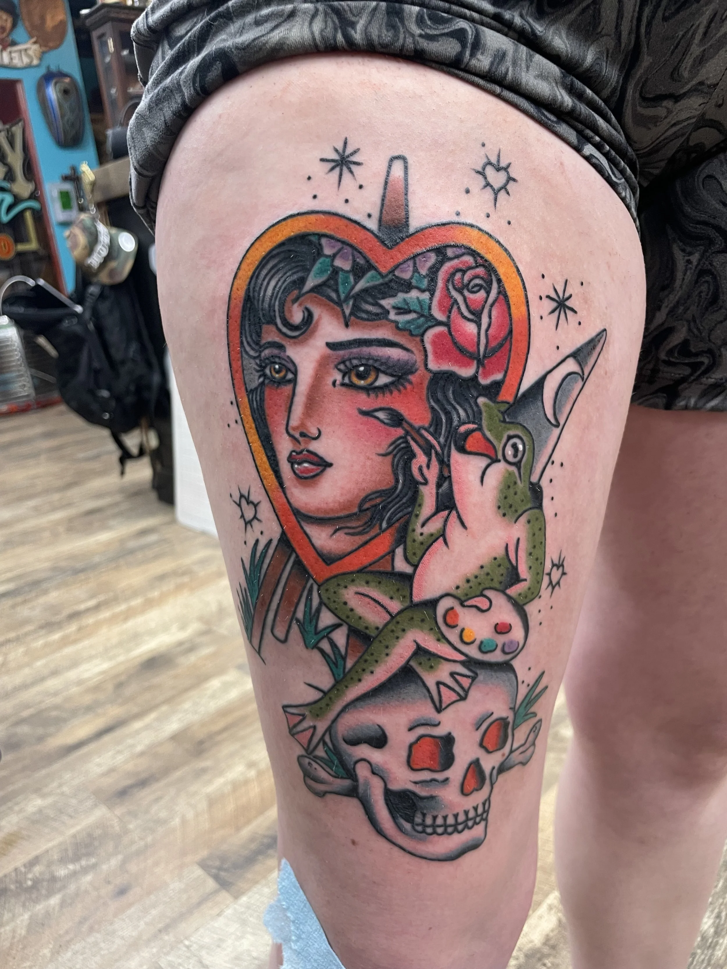 Colorful tattoo of a woman's face with a skull at the bottom, surrounded by stars, a heart, and a unicorn, on a person's thigh.