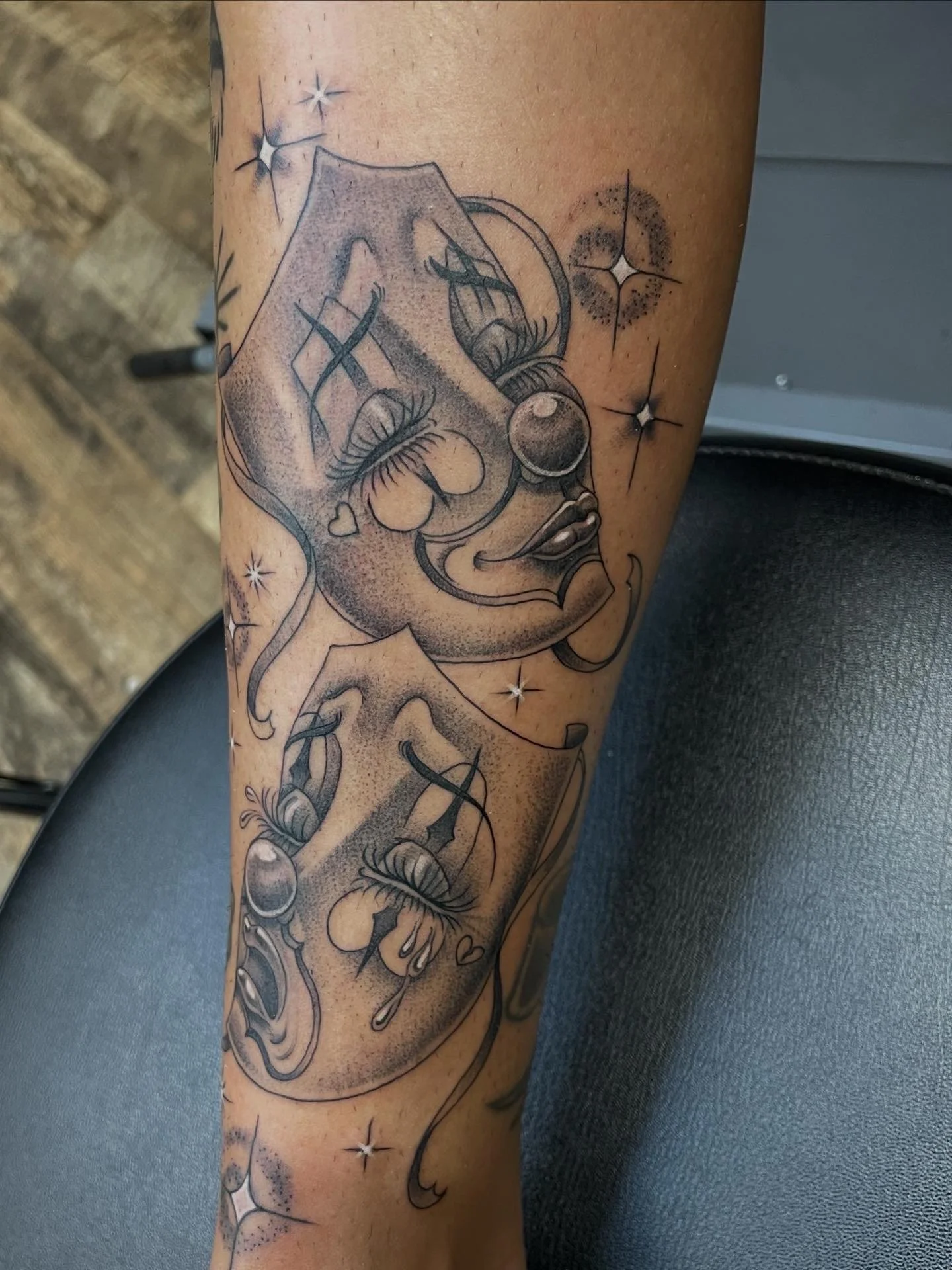Black and gray tattoo of a clown face with exaggerated features, styled with eyelashes, a nose ring, and a wide smile, surrounded by stars and heart symbols, on a person's arm.
