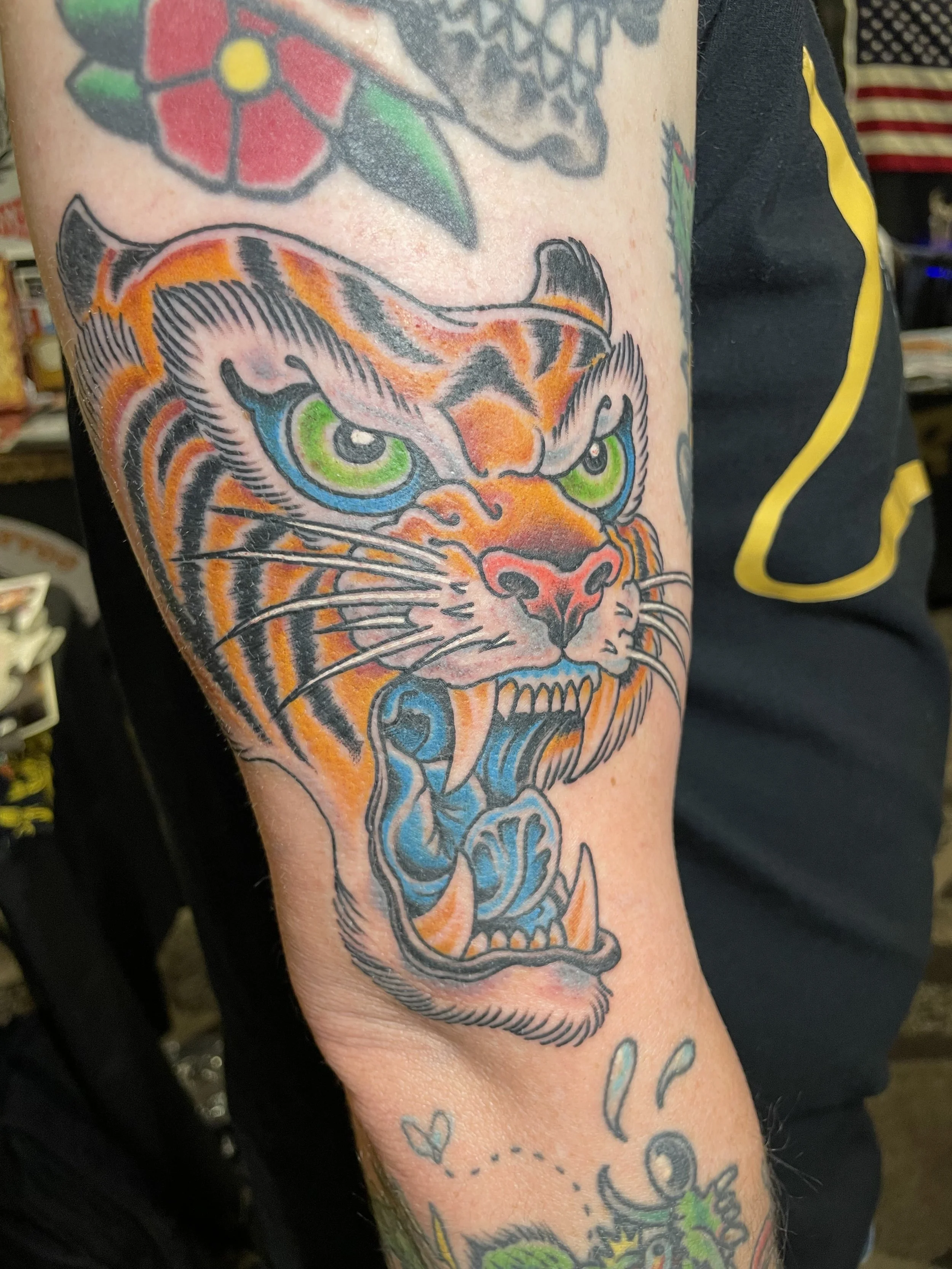 Close-up of a colorful traditional tattoo on a person's arm, featuring a fierce tiger's face with bright green eyes, orange and black stripes, and detailed facial features.