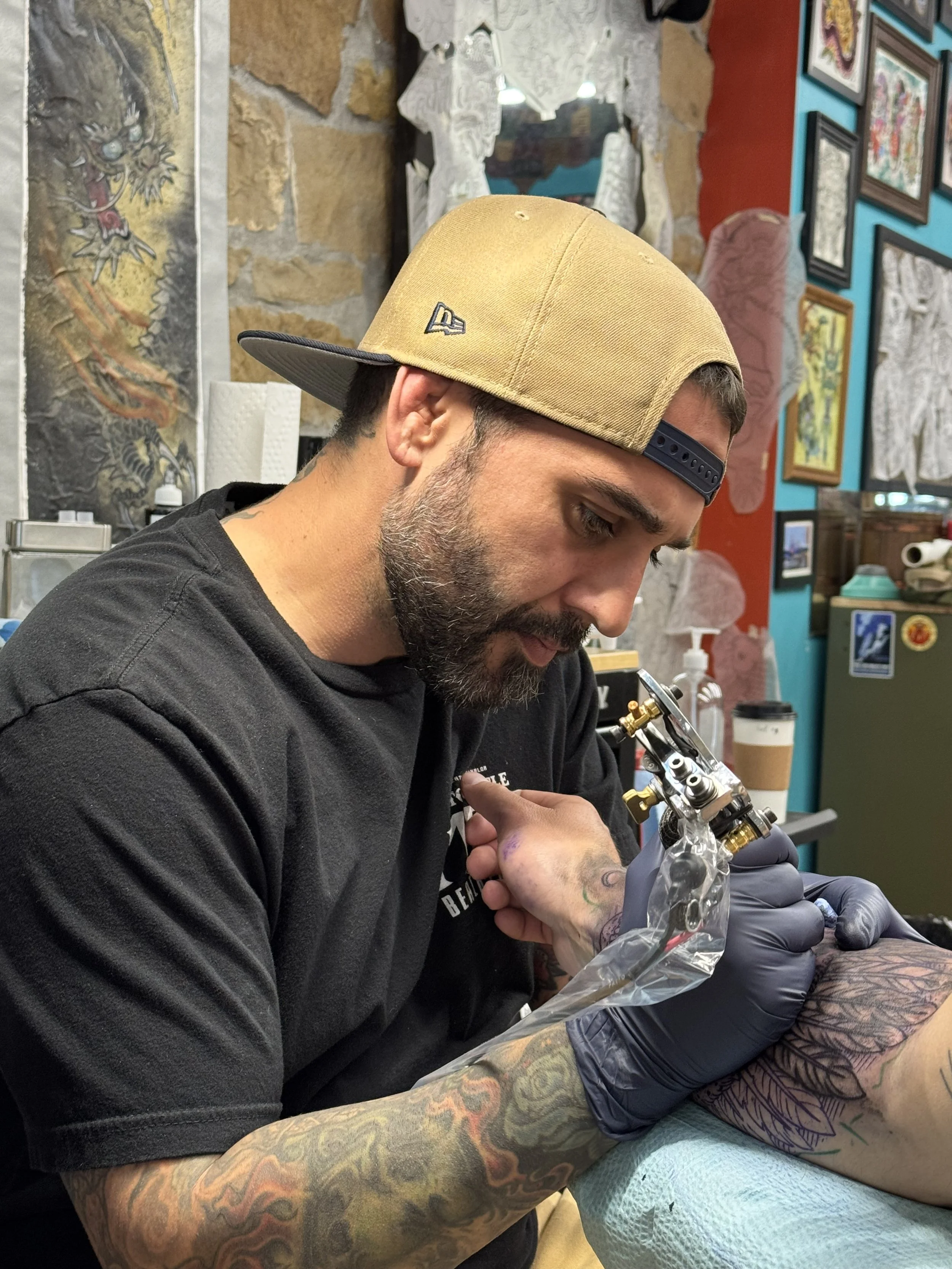 A man with a beige cap and a black shirt tattooing a person's leg in a tattoo studio.