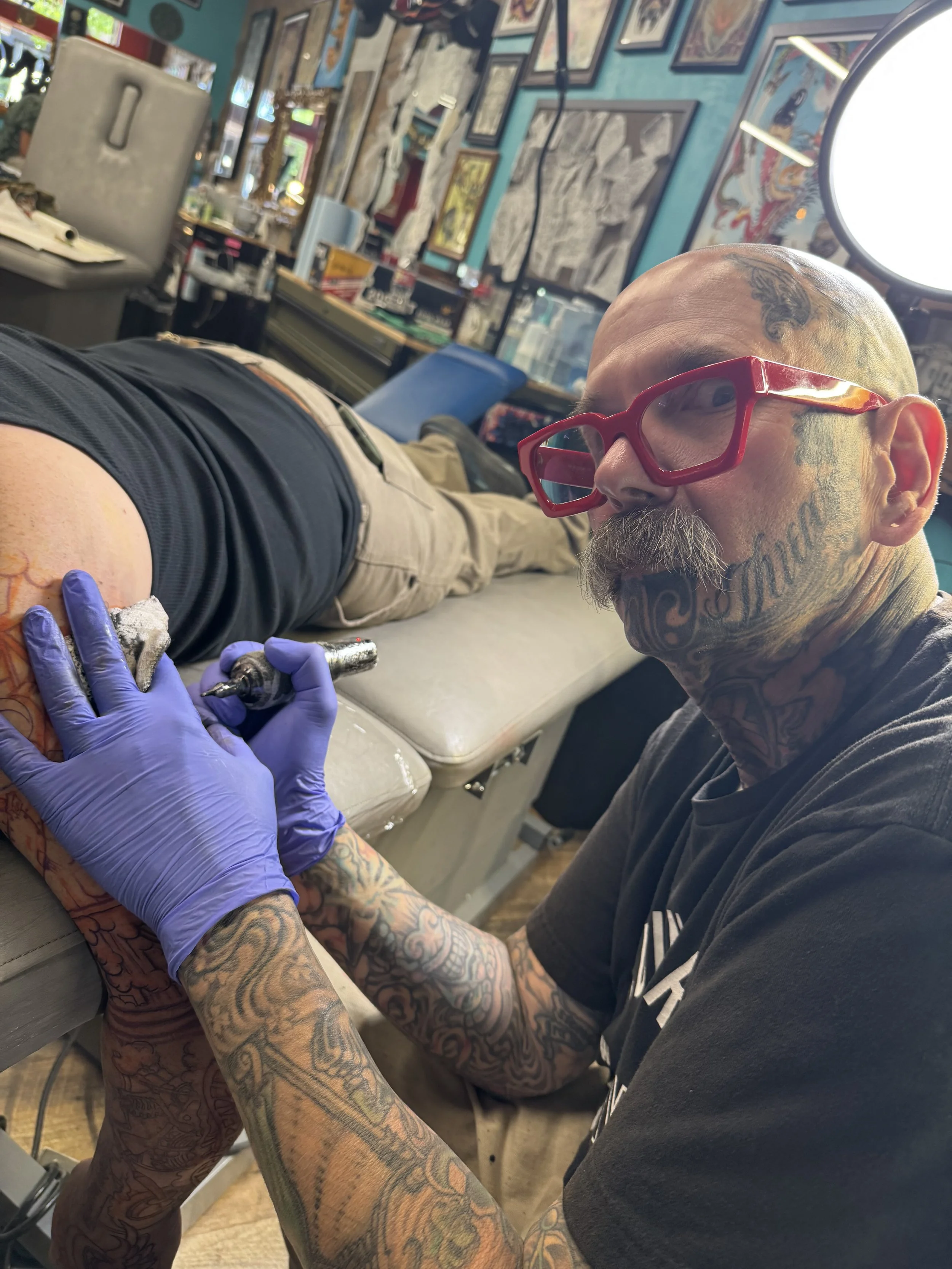 A tattooed man with glasses getting a tattoo on his arm by an artist wearing gloves in a tattoo studio decorated with framed artwork on the walls.