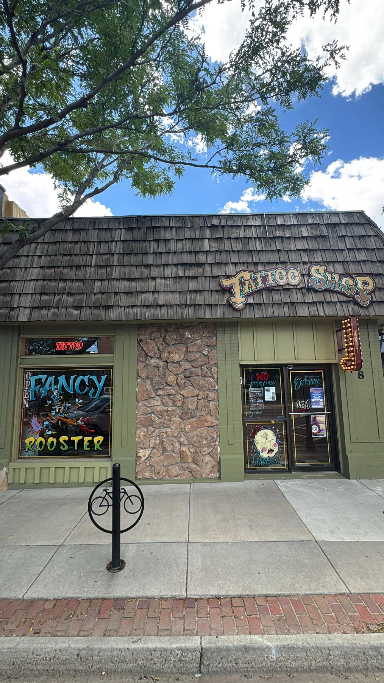 Front of a tattoo shop with large colorful signage and window artwork, featuring a stone accent wall and wooden shingle roof, trees, and a bright blue sky with white clouds.