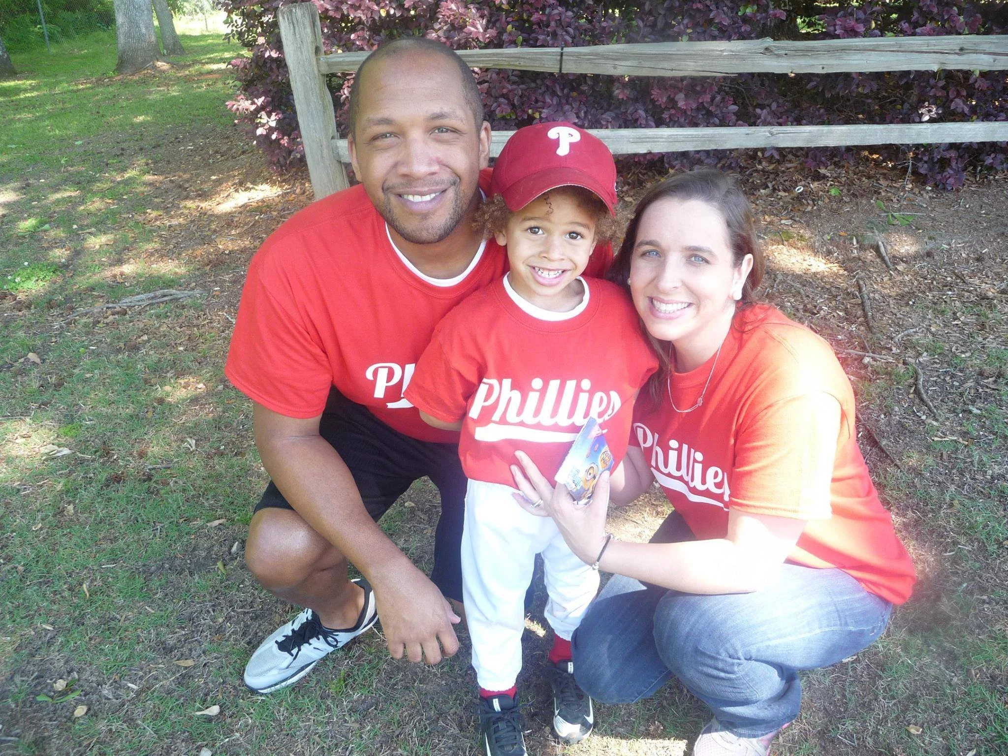 A smiling family of three sitting on the ground outside in a park, wearing matching red Phillies shirts. There is a man, a woman, and a young boy, with the boy wearing a red Phillies cap.