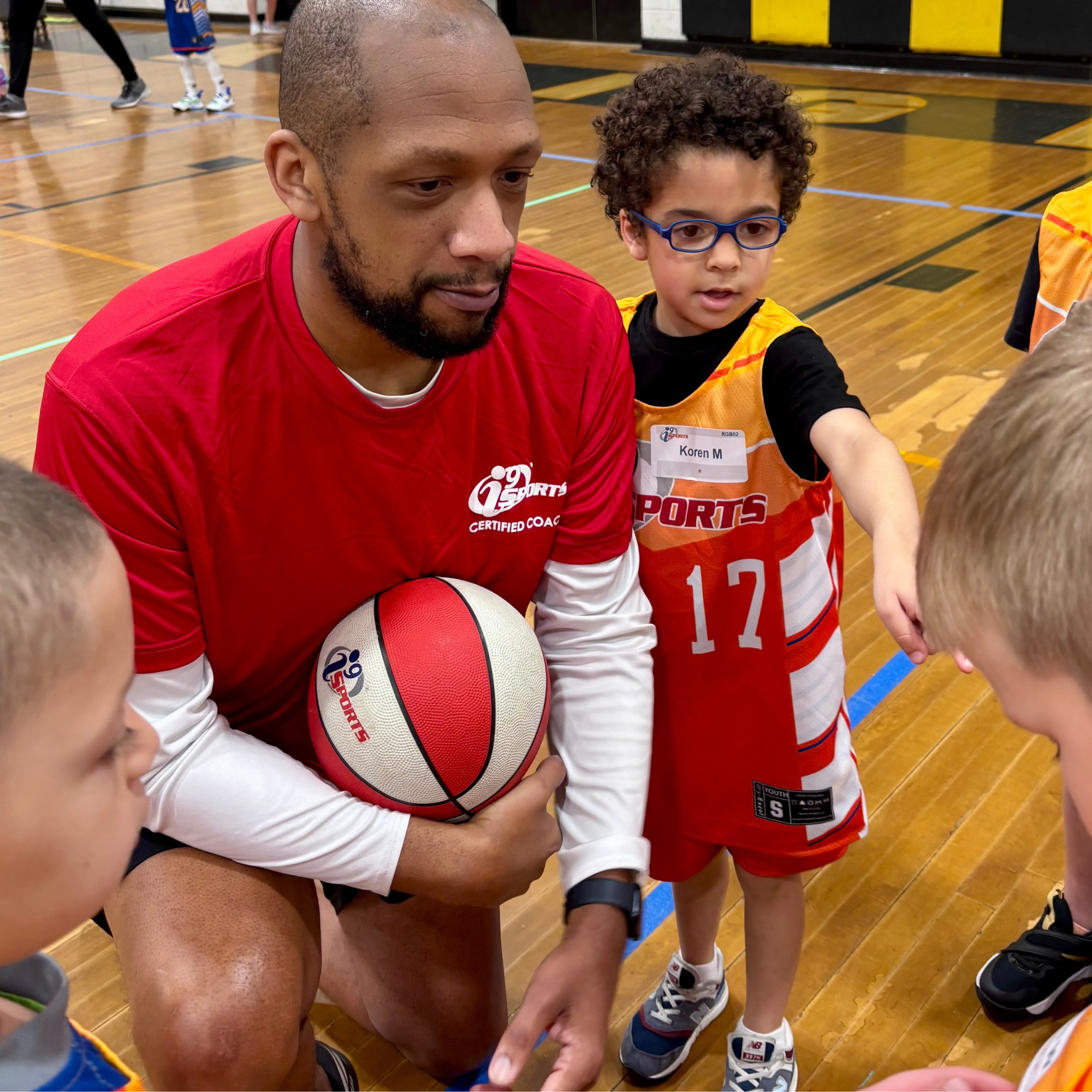 A man and a young boy sitting on the floor of a basketball gym, holding a basketball, surrounded by other children, with the man wearing a red sports shirt and the boy wearing a yellow sports jersey with number 17 and blue glasses.