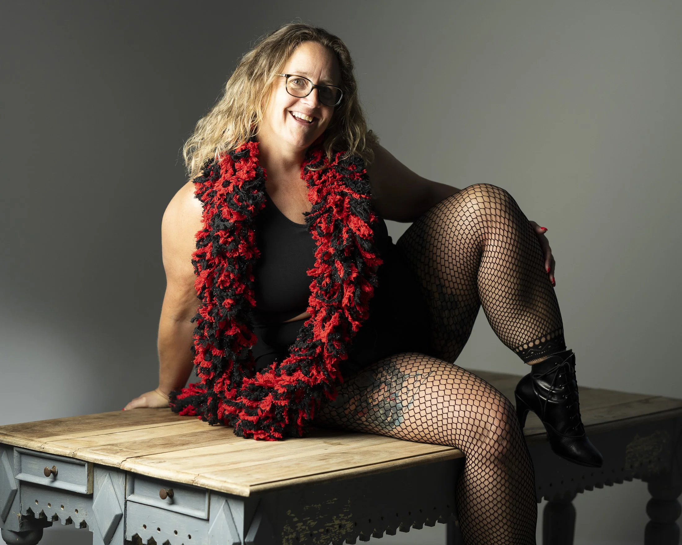 A woman with wavy hair, glasses, and a big smile sitting on a wooden table, wearing fishnet stockings, a black top, and a red and black knitted scarf, against a plain gray background.