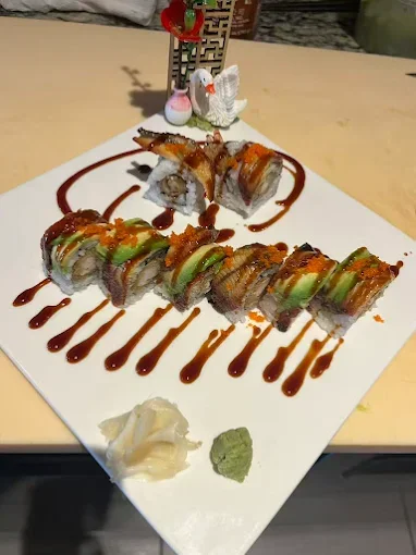 Plate of sushi rolls drizzled with sauce and served with wasabi and ginger, decorated with a small holiday-themed decoration and a white swan figurine.
