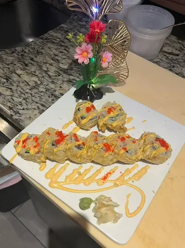 A sushi cake decorated with orange and red fish roe, with two decorative sushi roll pieces on top, served on a white square platter with wasabi and ginger, and pink and red artificial flowers in the background.