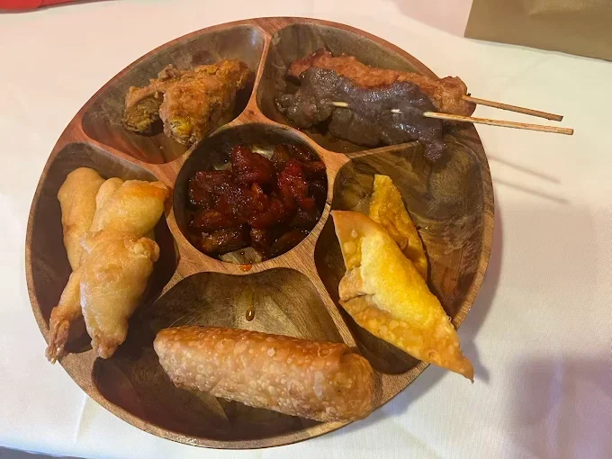 A wooden platter with seven sections containing different Asian appetizers, including spring roll, fried shrimp, satay skewers, fried chicken, fried wonton, a dark dipping sauce, and another fried item.