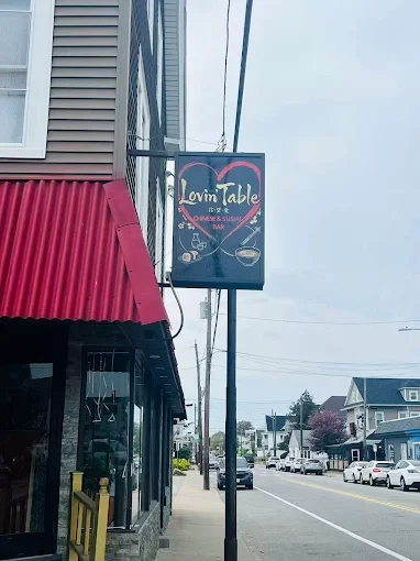 Sign for Lovin Table Chinese & Sushi Bar hanging on a street sidewalk next to a building with a red awning.
