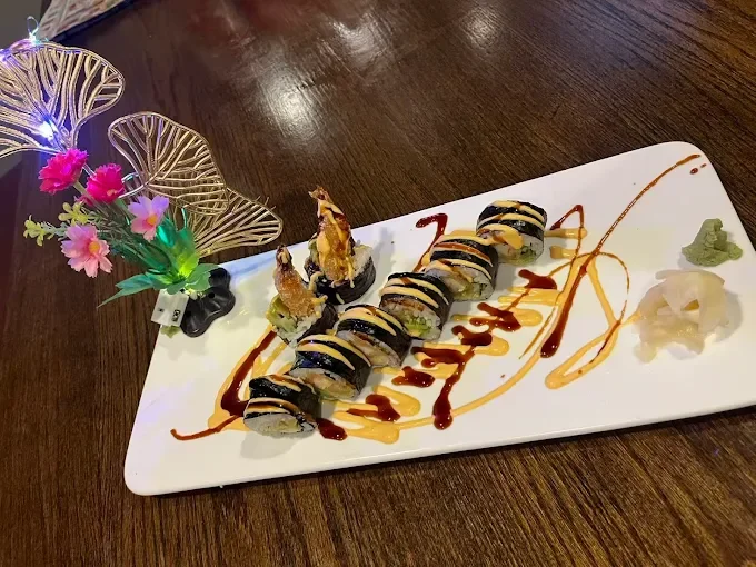 A sushi roll decorated with sauces, presented on a white rectangular plate, garnished with wasabi, ginger, and decorative flower and LED light arrangement.