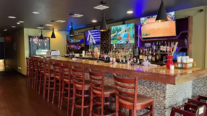 Empty bar with wooden counter and red chairs in a dimly lit restaurant or pub, with multiple television screens mounted on the wall showing sports, and various bottles and bar tools on the counter.