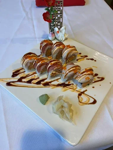 Sushi rolls drizzled with chocolate sauce, garnished with a cherry blossom decoration, wasabi, and pickled ginger on a white rectangular plate.