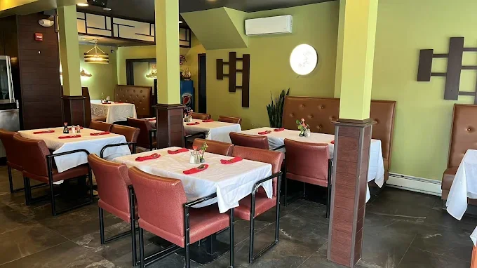 Restaurant dining area with tables covered in white tablecloths, red napkins, and small flower arrangements. Pink upholstered chairs surround the tables. Green walls with decorative black wall hangings and a small window with a round light on the wal