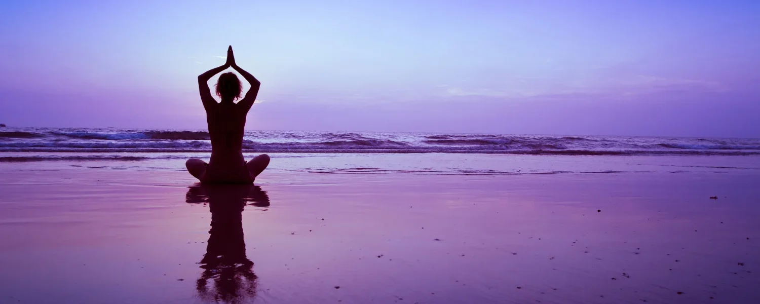Person practicing yoga in a seated meditation pose on a beach at sunset or sunrise.