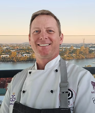 Male chef in a white chef coat and apron smiling outdoors with a cityscape and river in the background.