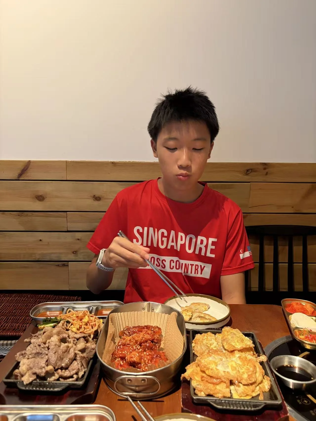 A young boy sitting at a table in a restaurant, wearing a red T-shirt with the word "SINGAPORE" printed on it, eating Korean food with chopsticks surrounded by various dishes.