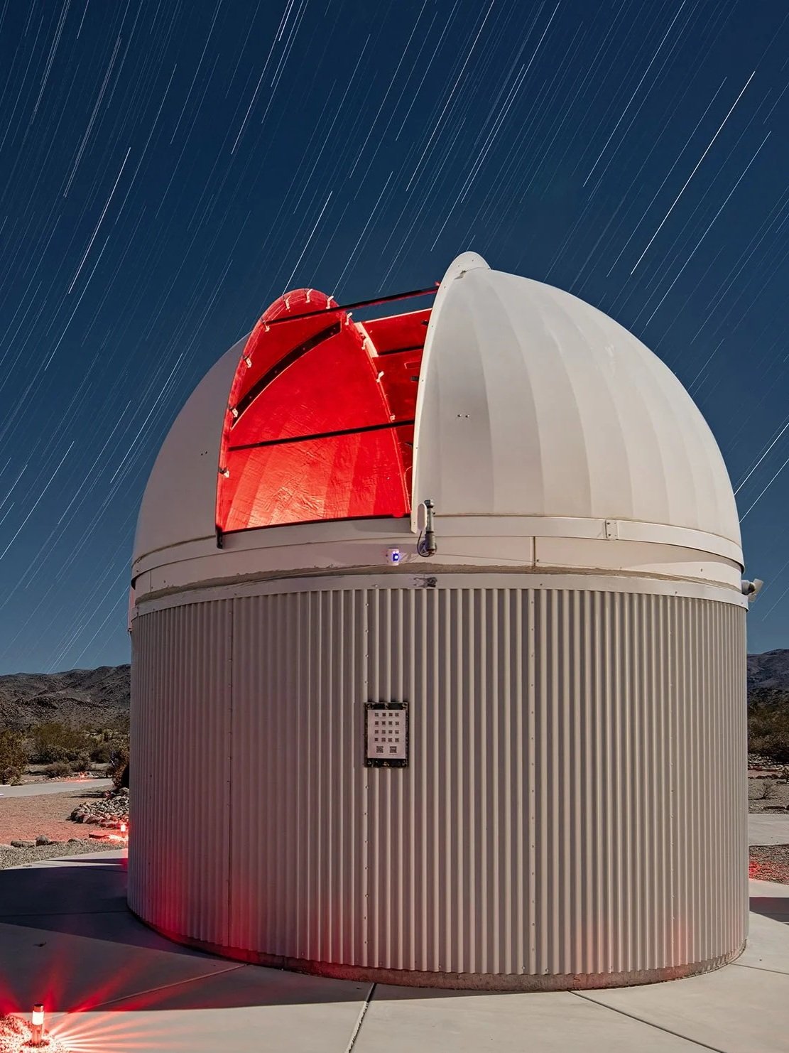 A large white observatory with its roof partially open, revealing a red interior, against a night sky with star trails.