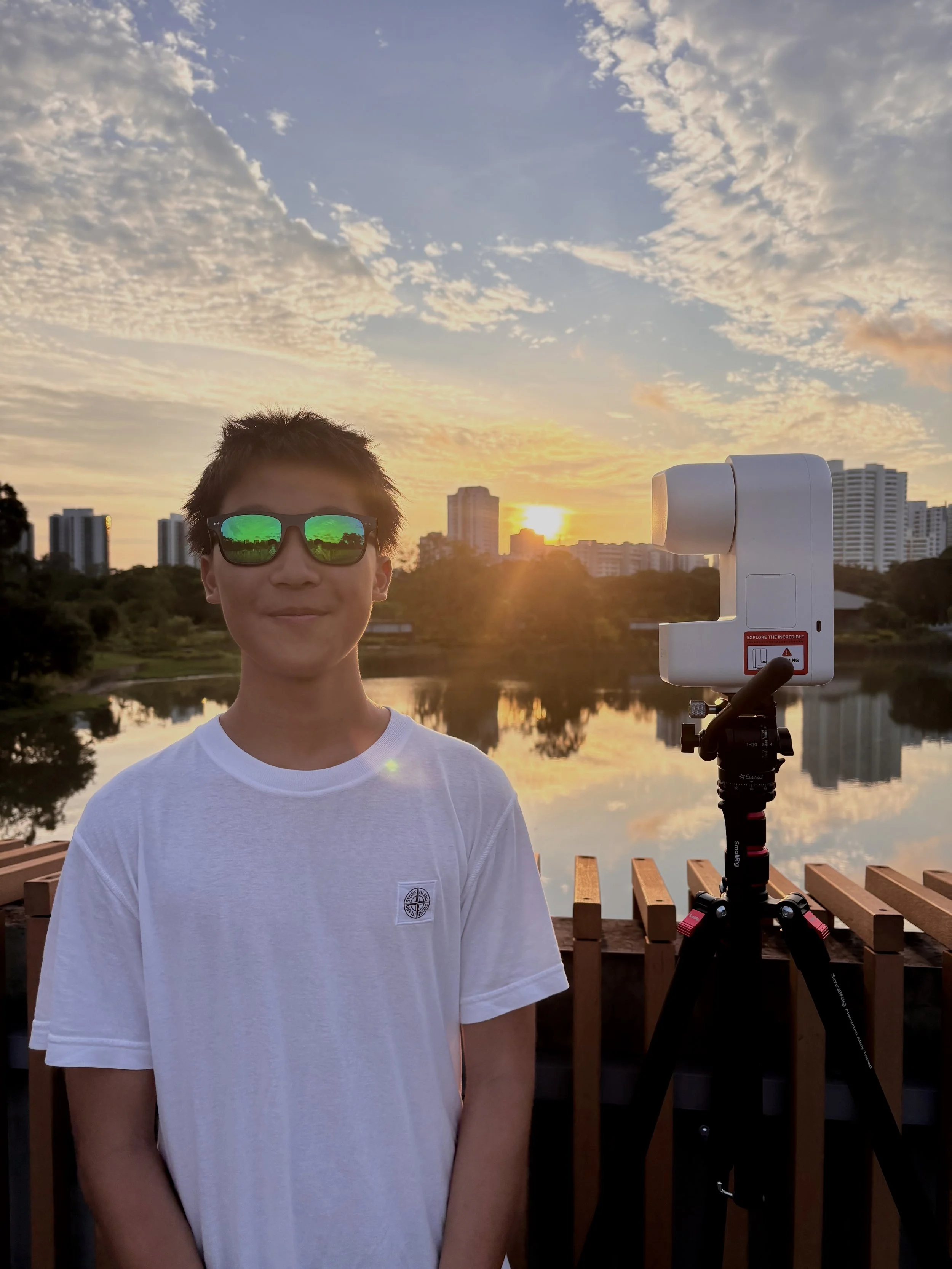 A young person with short, dark hair wearing reflective sunglasses and a white t-shirt standing outdoors during sunset, with a camera on a tripod to their right and city buildings in the background.