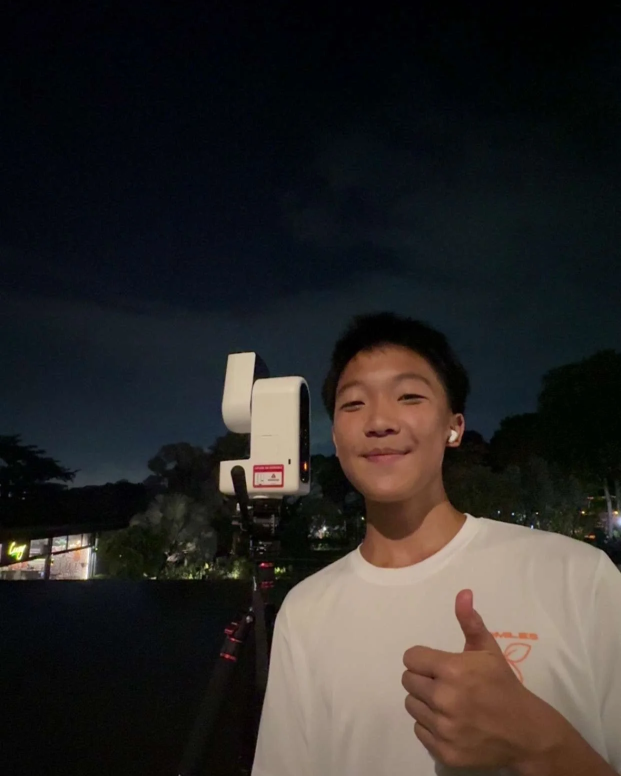 Young boy giving a thumbs-up in front of a surveyor instrument at night, with dark sky and trees in the background.