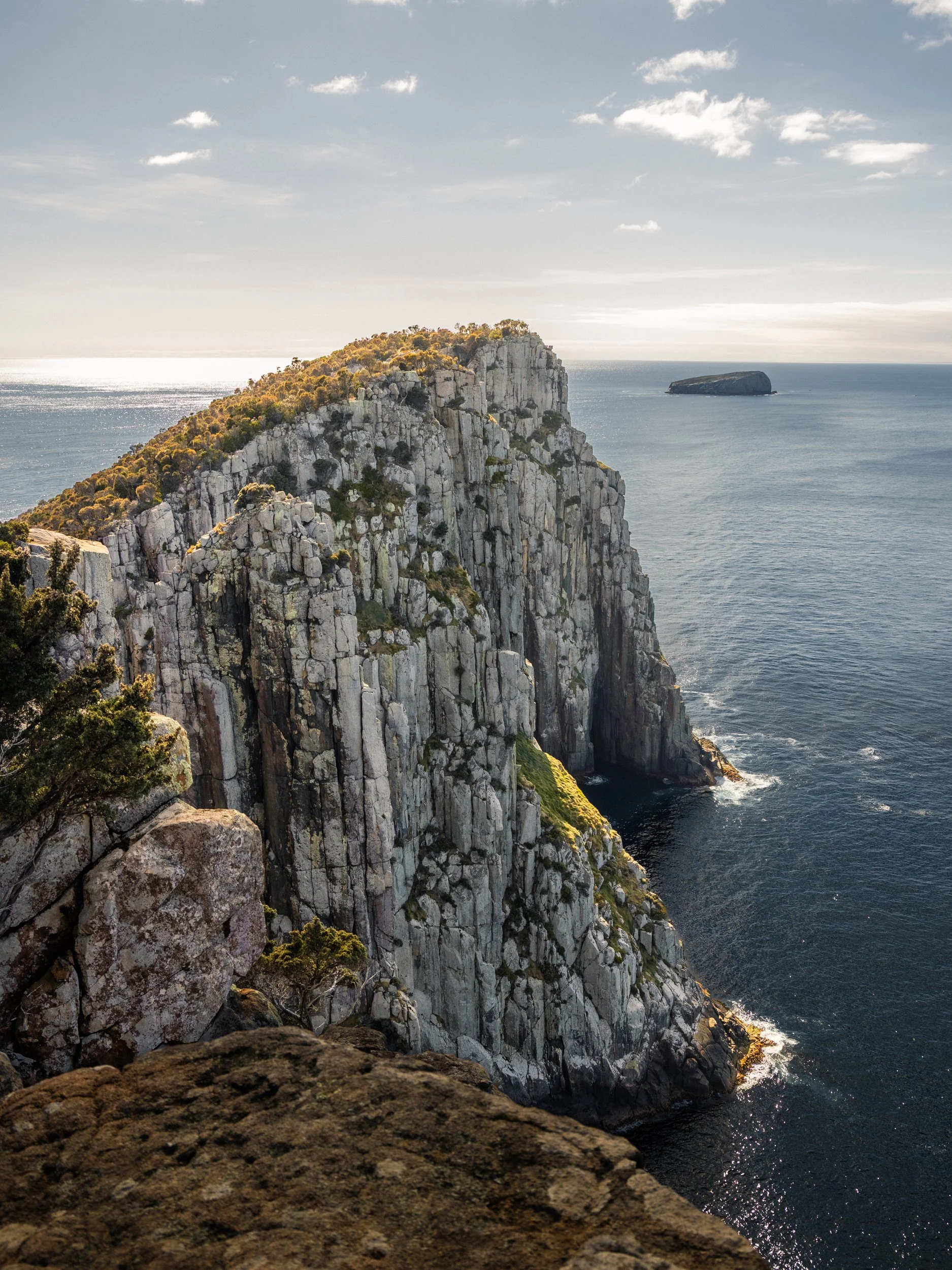 View of a rugged cliffside with white rocks and green vegetation overlooking the ocean, with a large rock formation in the water in the background under a partly cloudy sky.
