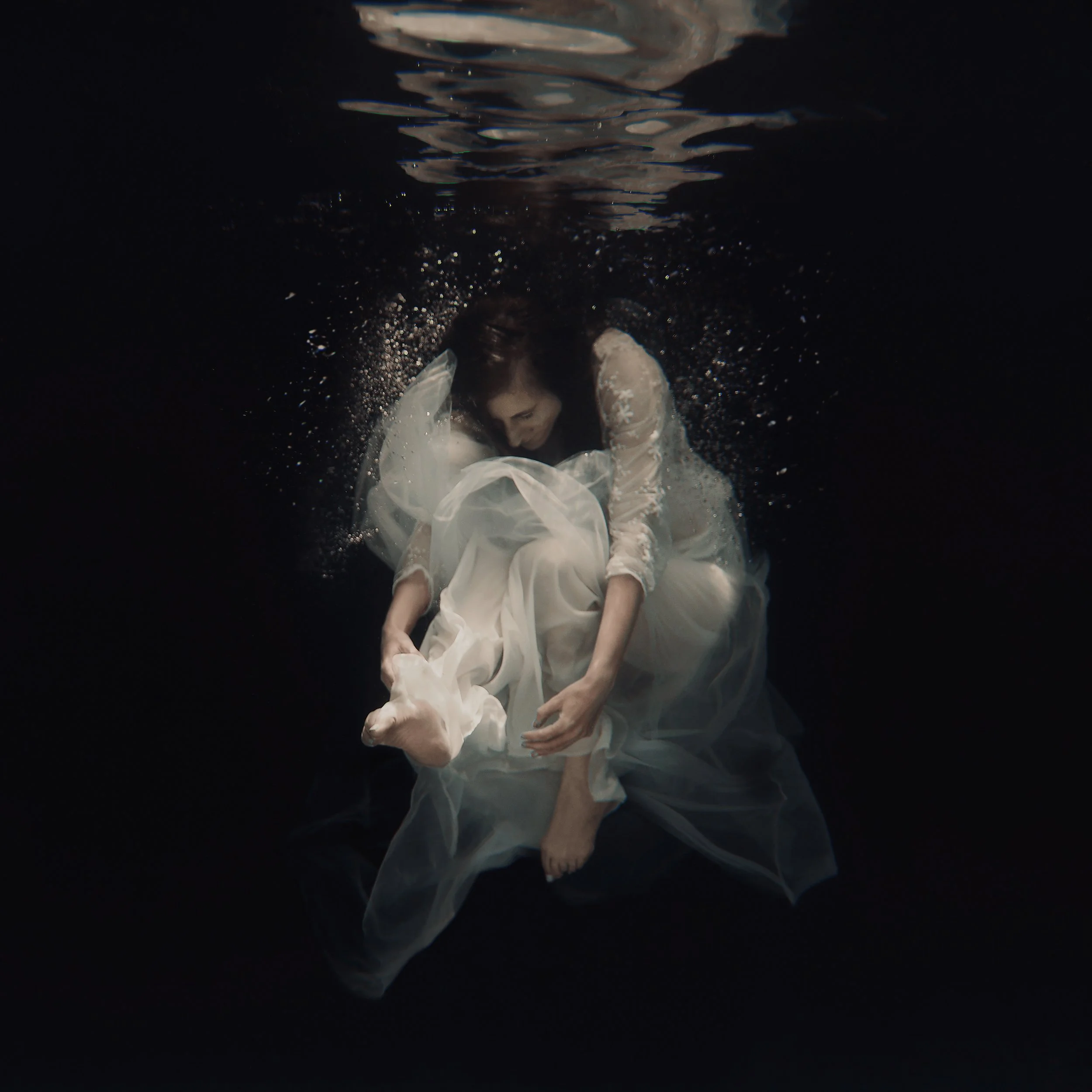 A person in a white dress or gown is underwater, with their head turned downward and with dark hair floating around. Their arms are bent at the elbows, and they are holding the fabric of the dress. The scene is dark with reflections on the water surf
