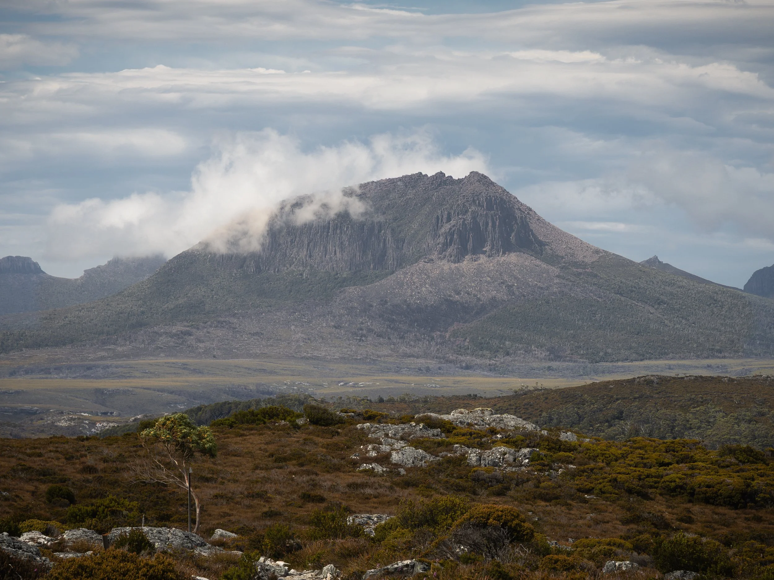 A mountain with dark, rugged slopes partially covered by clouds, with a foreground of rocky and bushy terrain under a cloudy sky.