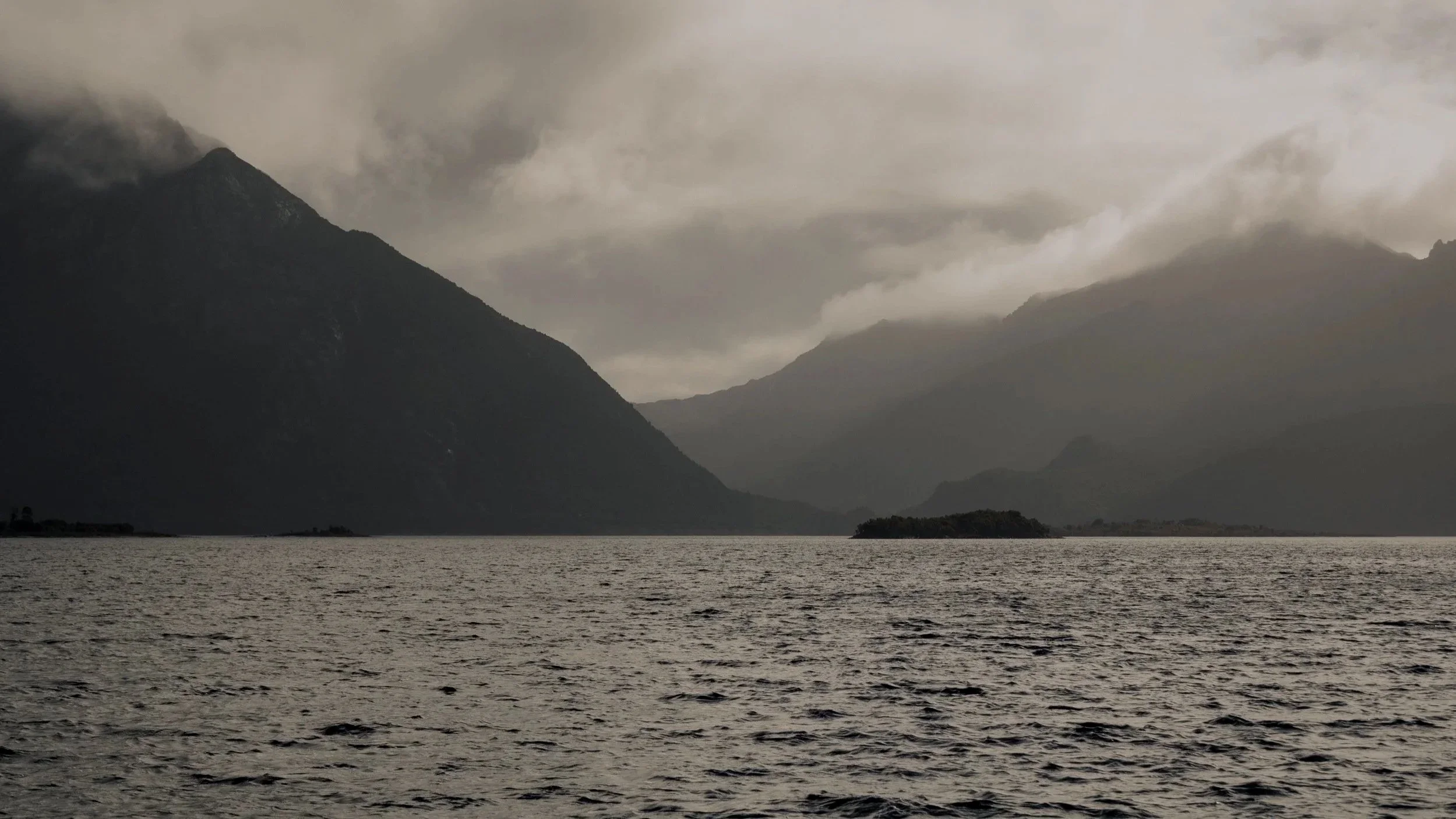 Dark and cloudy mountains overlooking a large body of water, with an overcast sky and mist or fog covering the mountain peaks.