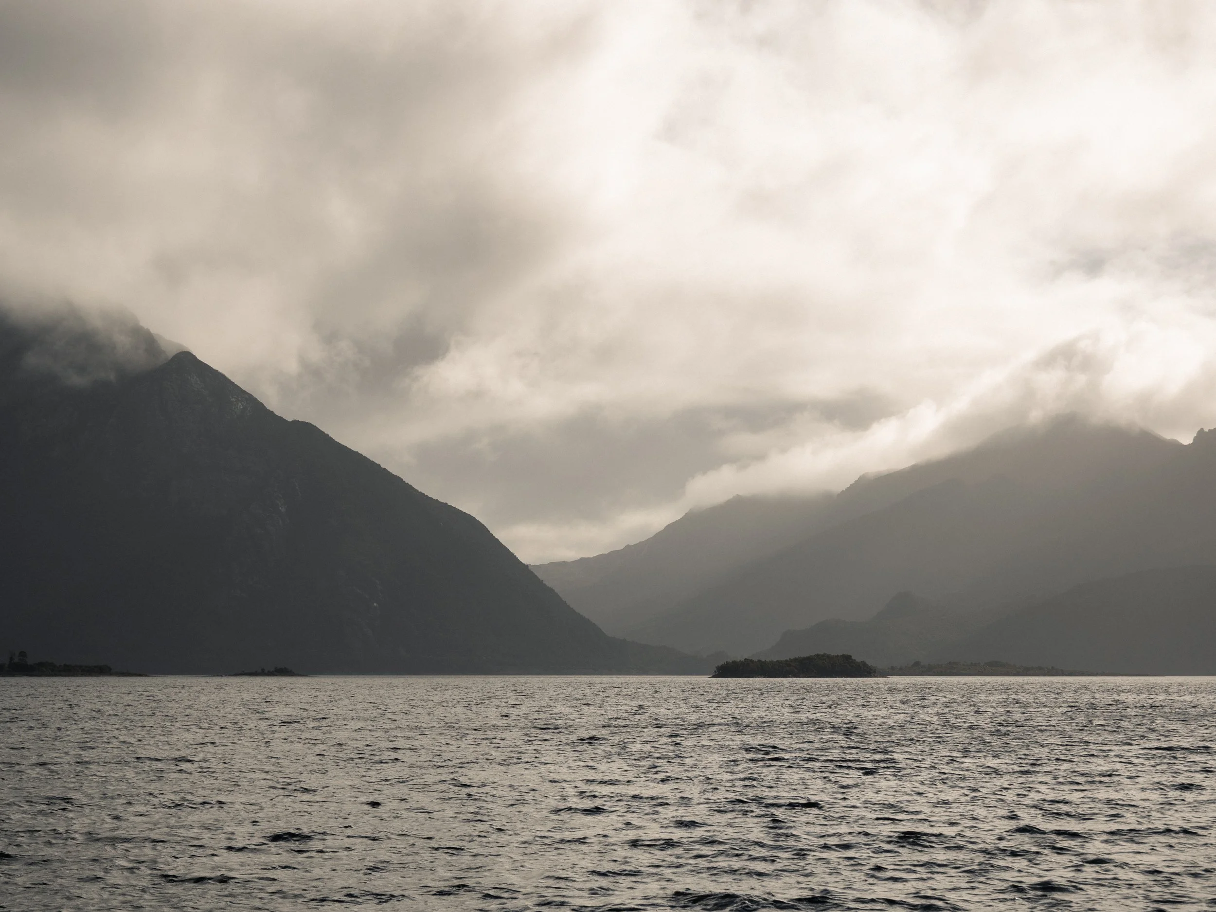 A mountain landscape with dark, cloudy sky over a body of water and a small island in the distance.