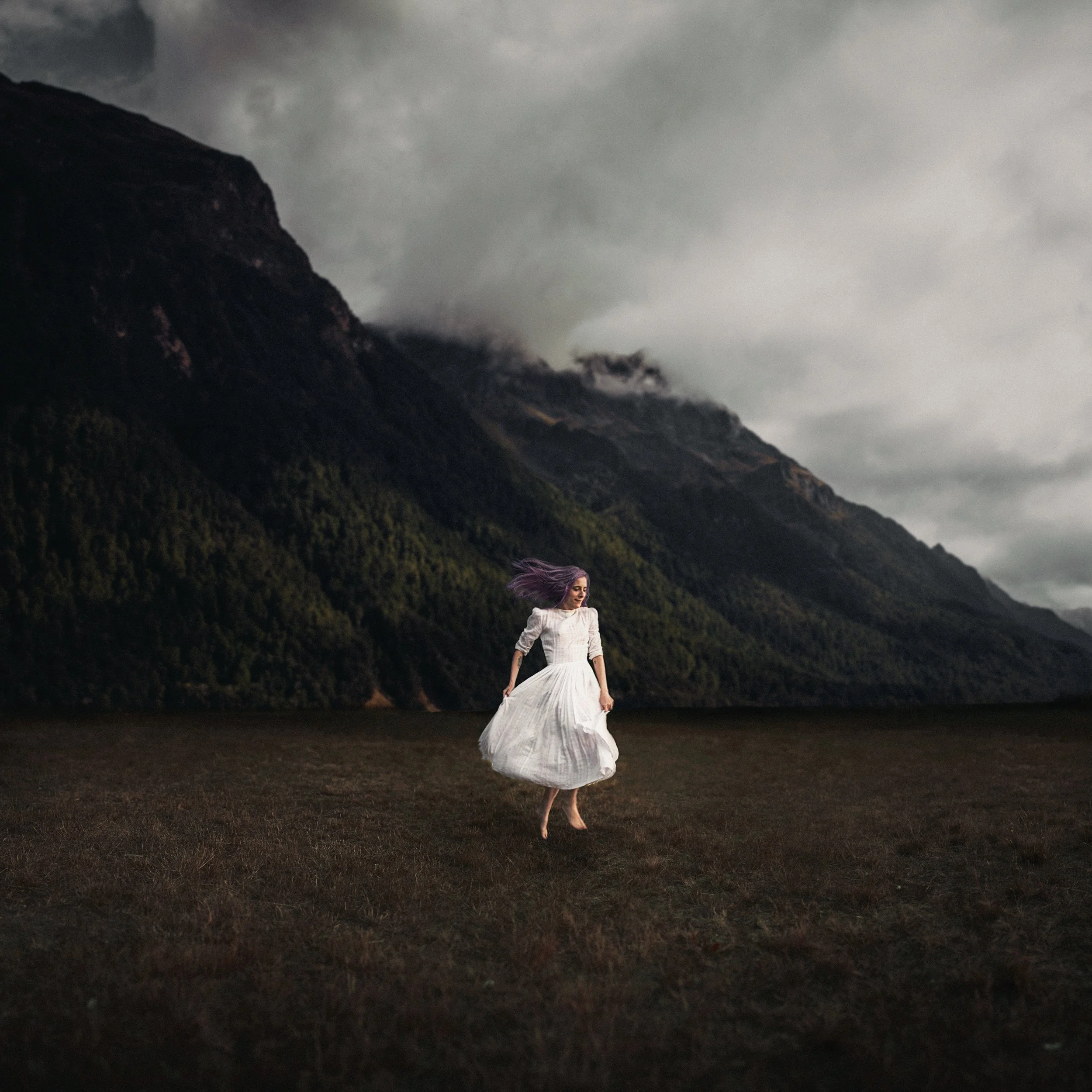 A woman in a white dress walking barefoot on grassy terrain with mountain and cloudy sky background.