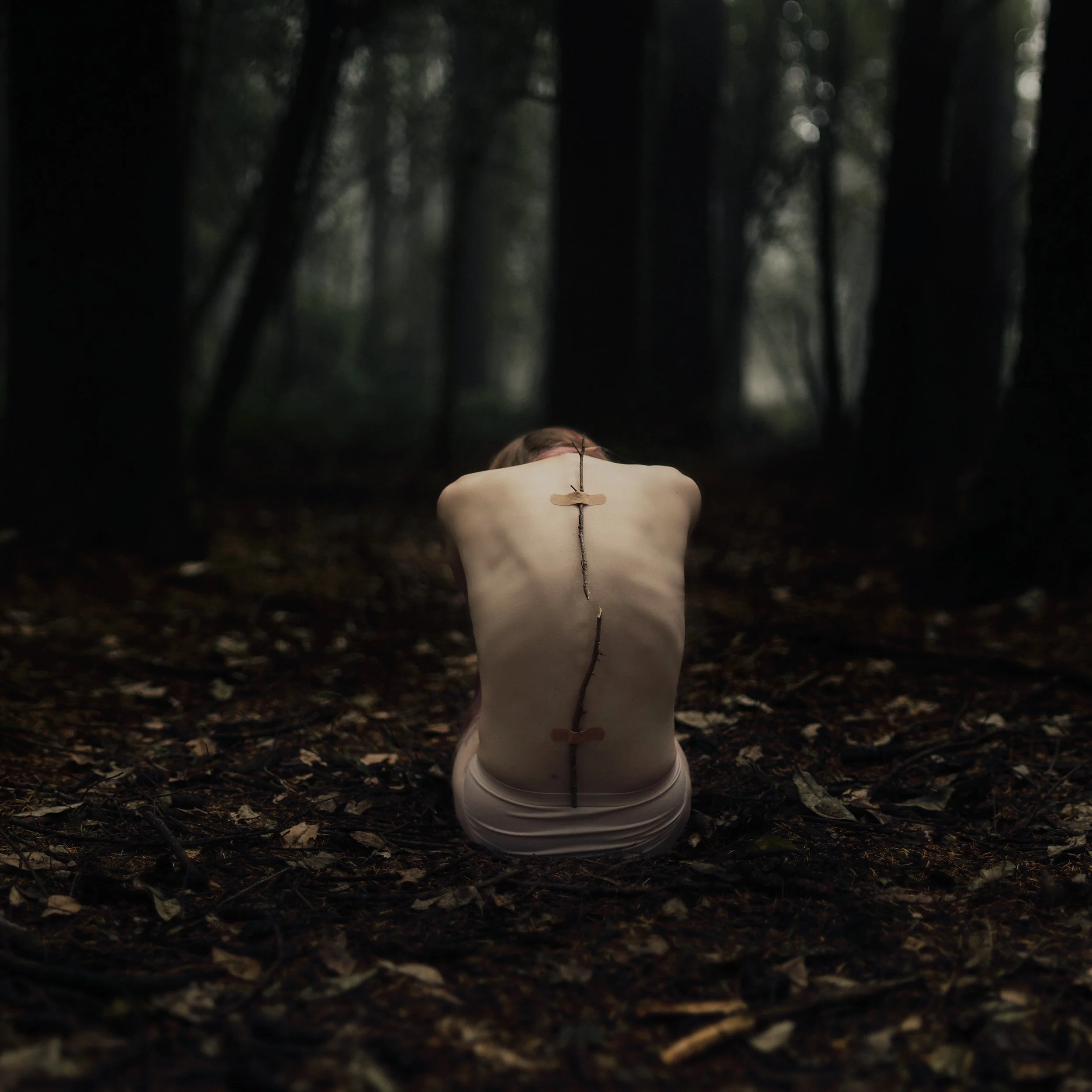 A mannequin lying face down on the forest floor in a dark wooded area, with a small branch on the back of its neck, and a cloth tied around its waist.