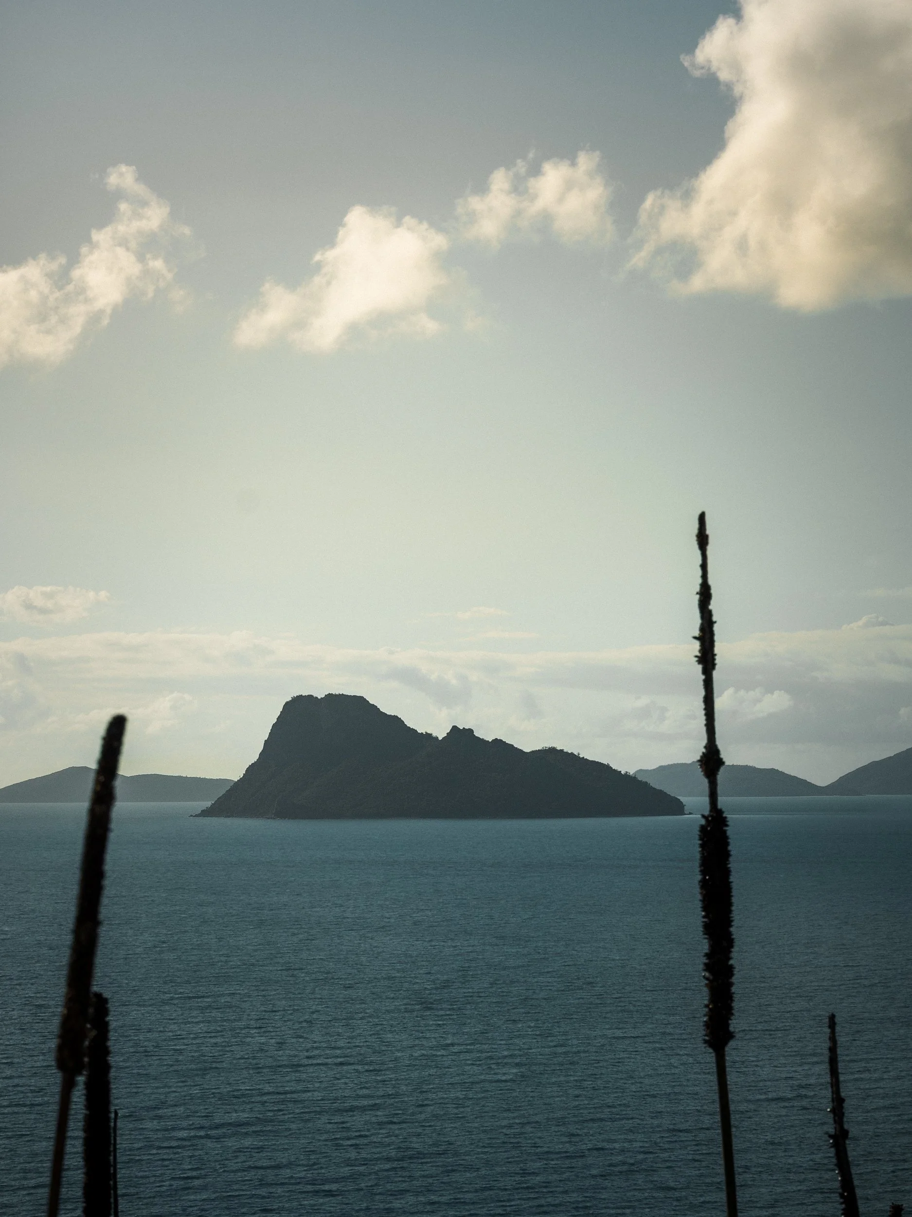 A scenic view of a calm body of water with an island in the distance, a mountain behind it, and a partially cloudy sky above. Foreground features two vertical sticks.