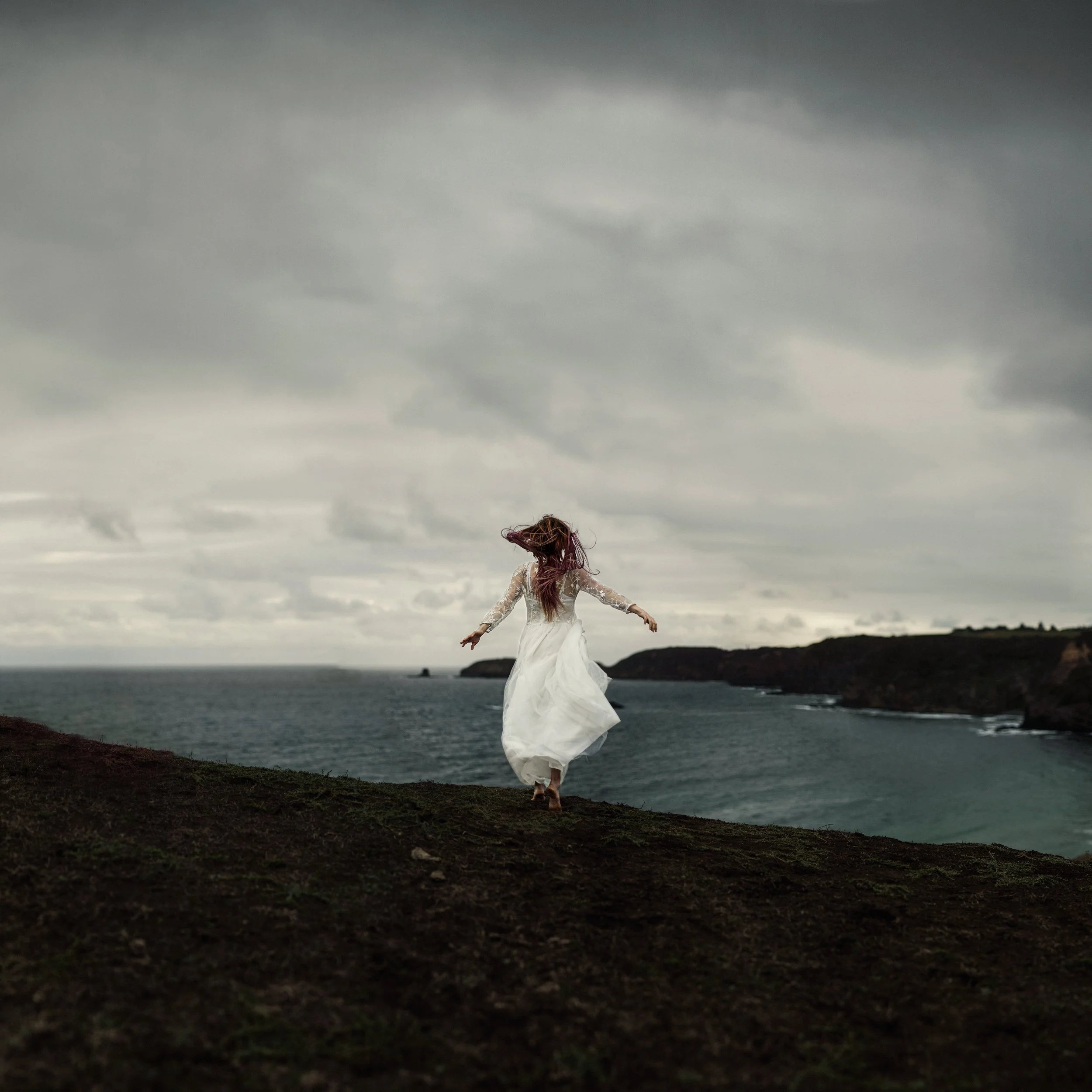 A person in a white dress with long hair standing on a rocky coastline with a body of water and a cloudy sky in the background.