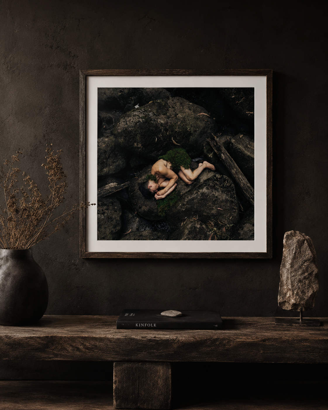 Framed photograph of a person curled up among moss-covered rocks in a natural setting, displayed on a dark textured wall above a rustic wooden shelf.