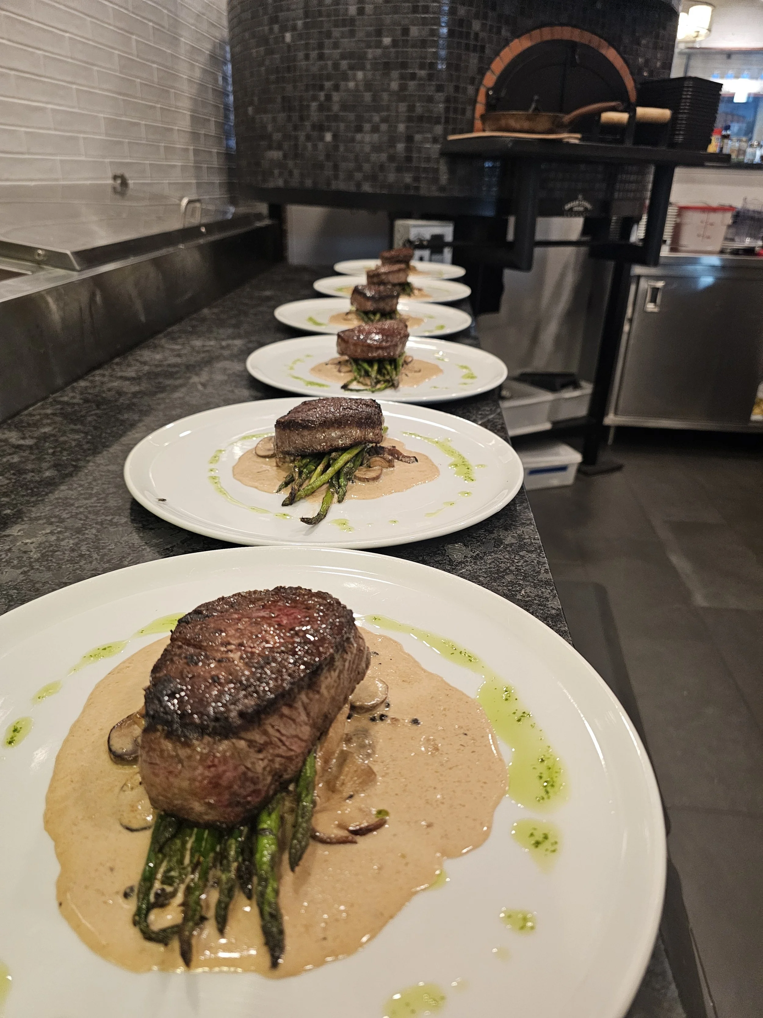 Multiple white plates of a cooked beef steak with green beans, mushrooms, and sauce, garnished with green sauce drizzled around the plates, set on a black countertop in a restaurant kitchen.