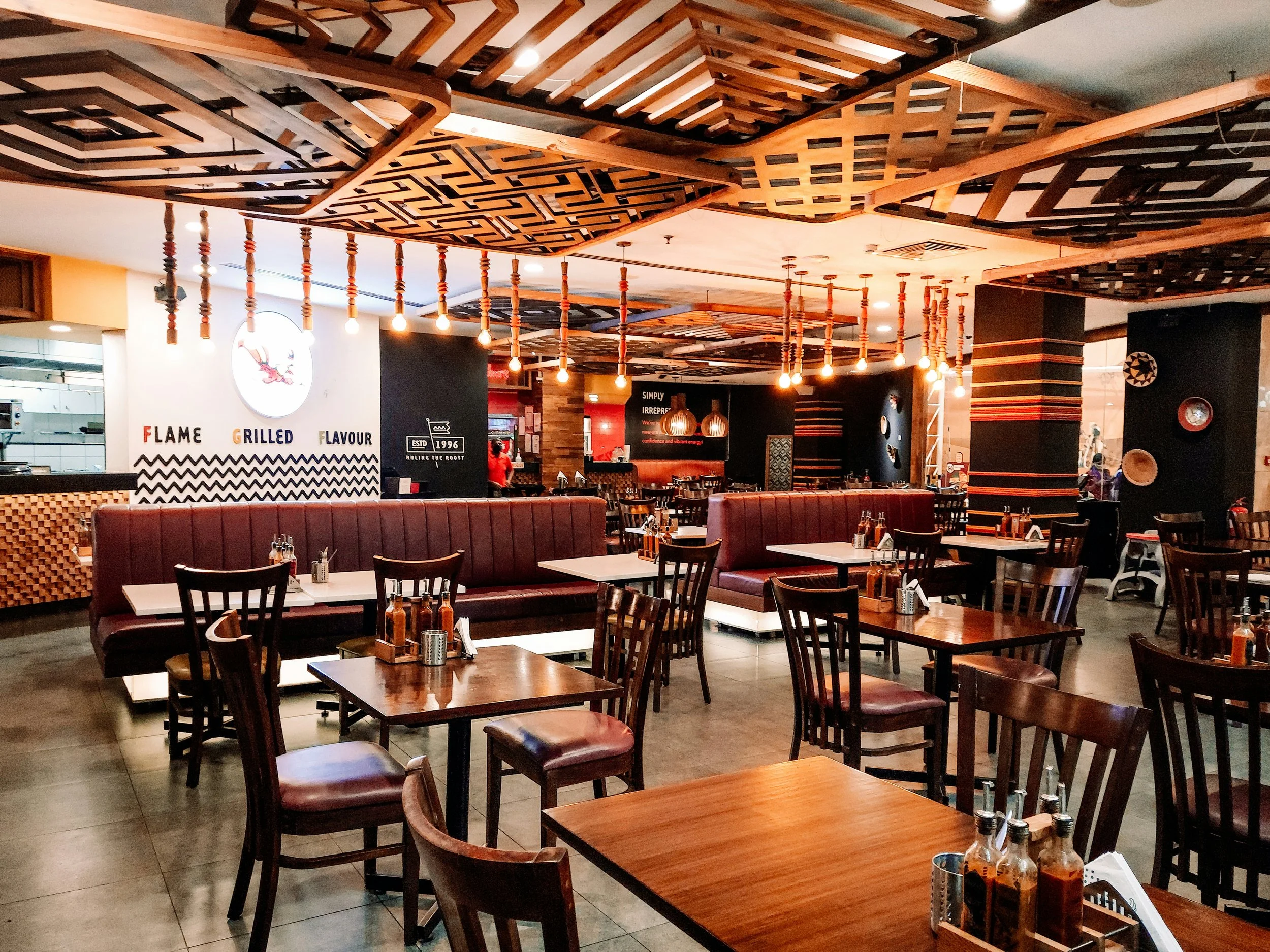 Interior of a restaurant with wooden tables and chairs, red booth seating, decorative wooden ceiling panels, hanging light bulbs, and black accent walls with art and signage.