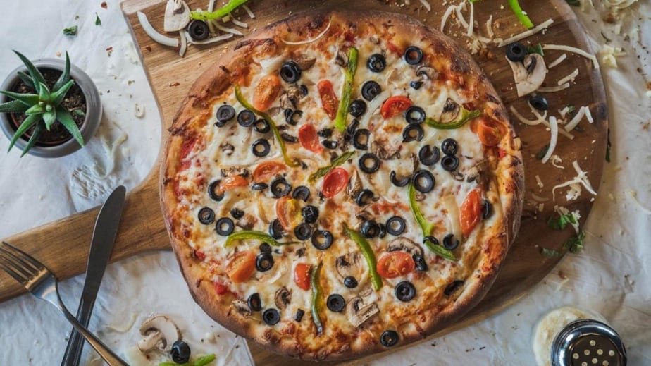 A pizza topped with black olives, cherry tomatoes, green peppers, and mushrooms on a round wooden board. Surrounding the pizza are a small potted succulent plant, a fork and knife, some sliced onions, and a small dish of dipping sauce.