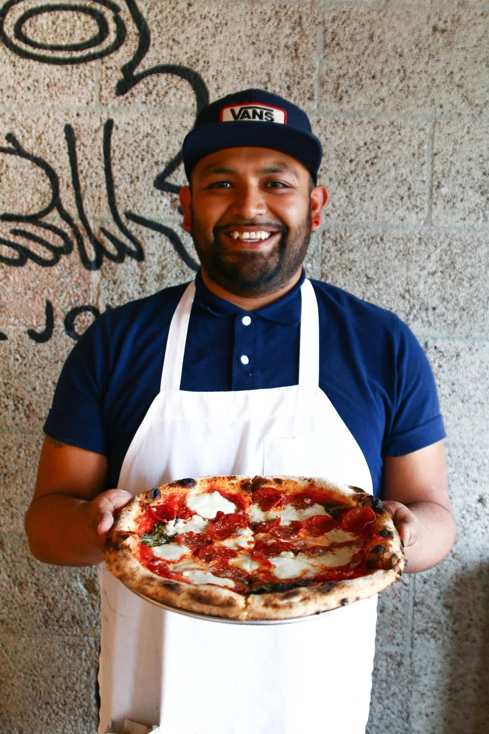 A smiling man wearing a navy blue cap with a Vans logo, a navy polo shirt, and a white apron, holding a pizza with tomato sauce, mozzarella cheese, and pepperoni, standing in front of a concrete wall with black graffiti.
