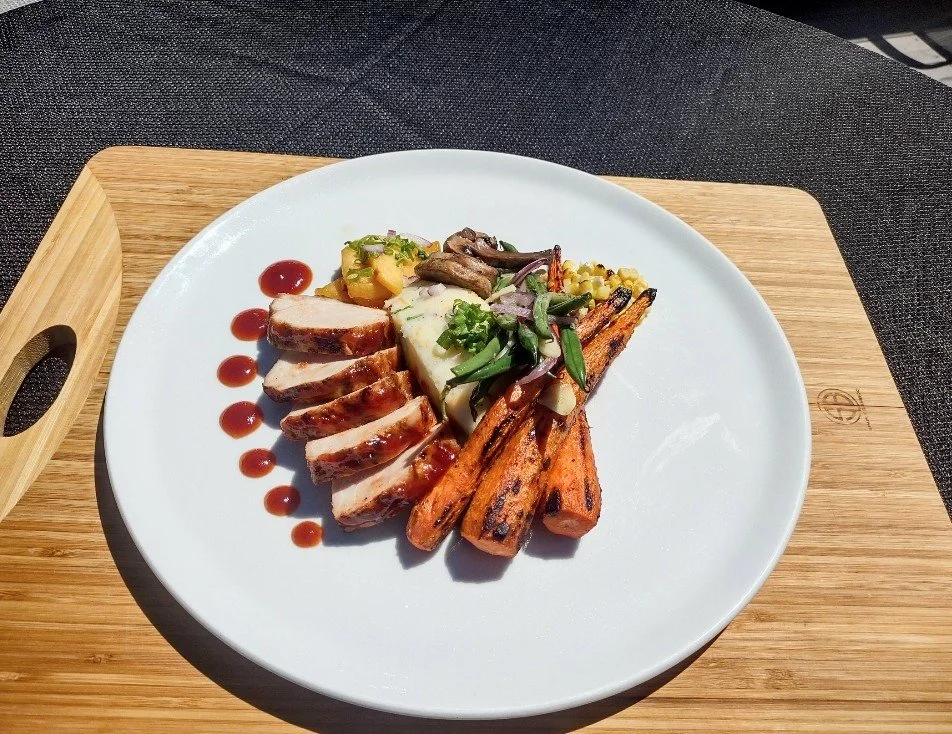 Plate with sliced grilled chicken, roasted carrots, grilled vegetables, and a dollop of mashed potato with chopped herbs, garnished with a small amount of sauce on a wooden tray.