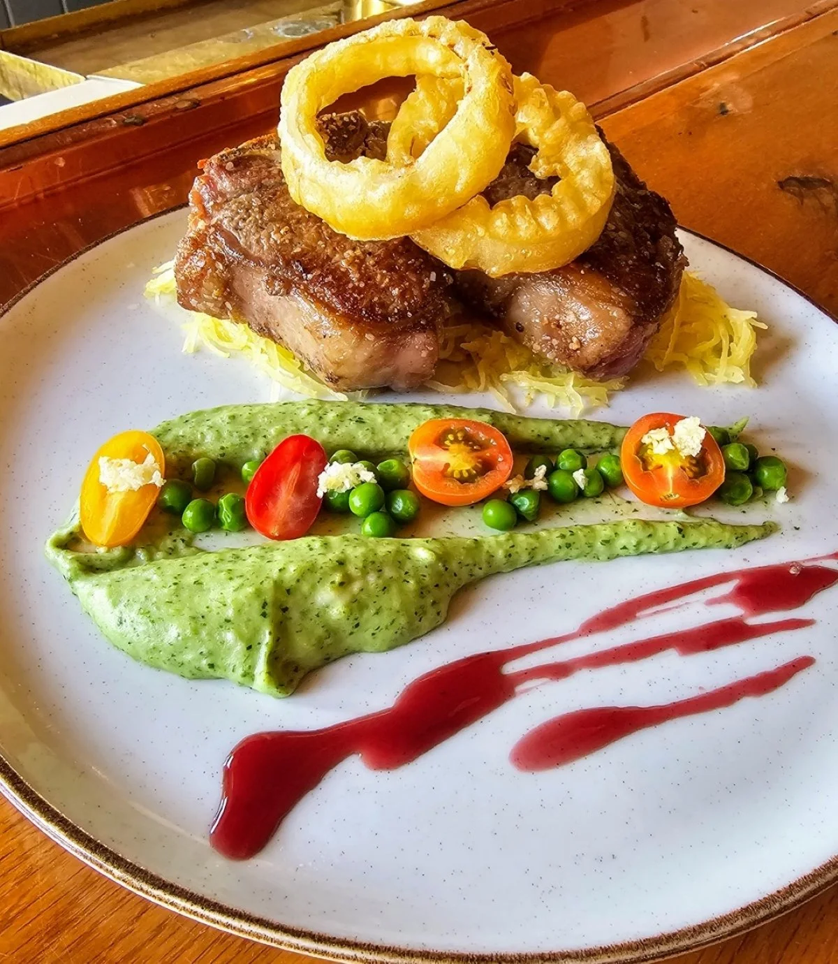 A plate featuring a grilled pork chop topped with onion rings, served with mashed potatoes and shredded lettuce. The plate also has a green pea puree with cherry tomato halves, green peas, and an herb drizzle, along with a decorative streak of red sa