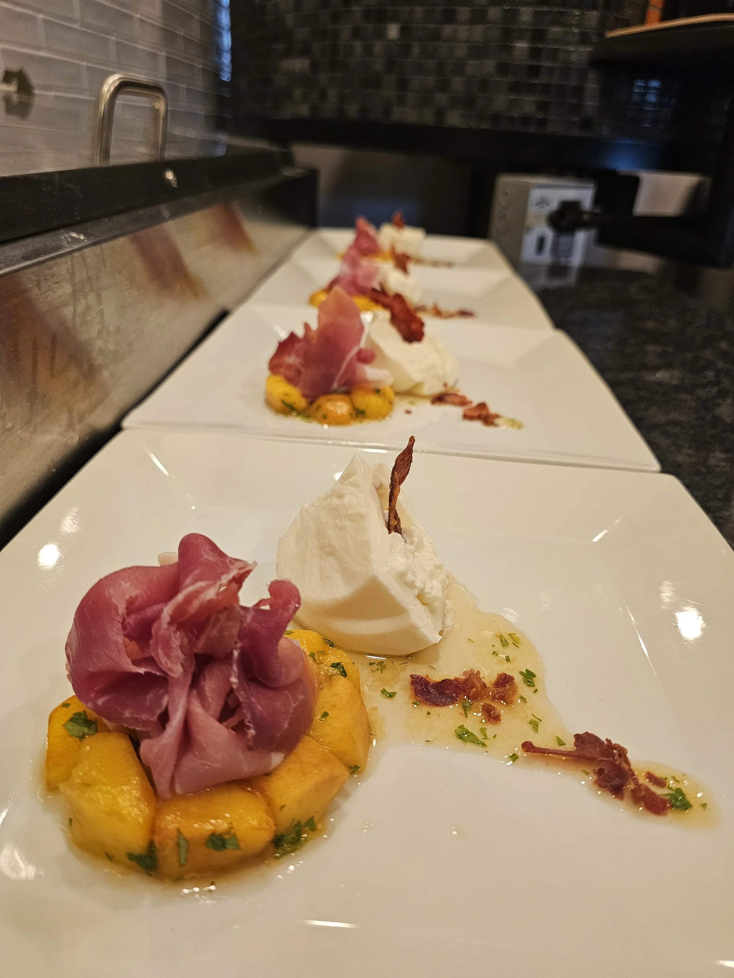 A close-up of a row of gourmet appetizers presented on white plates. The appetizers include slices of cured meats, vegetables, and garnishes, arranged with colorful sauces and herbs in a fine dining setting.