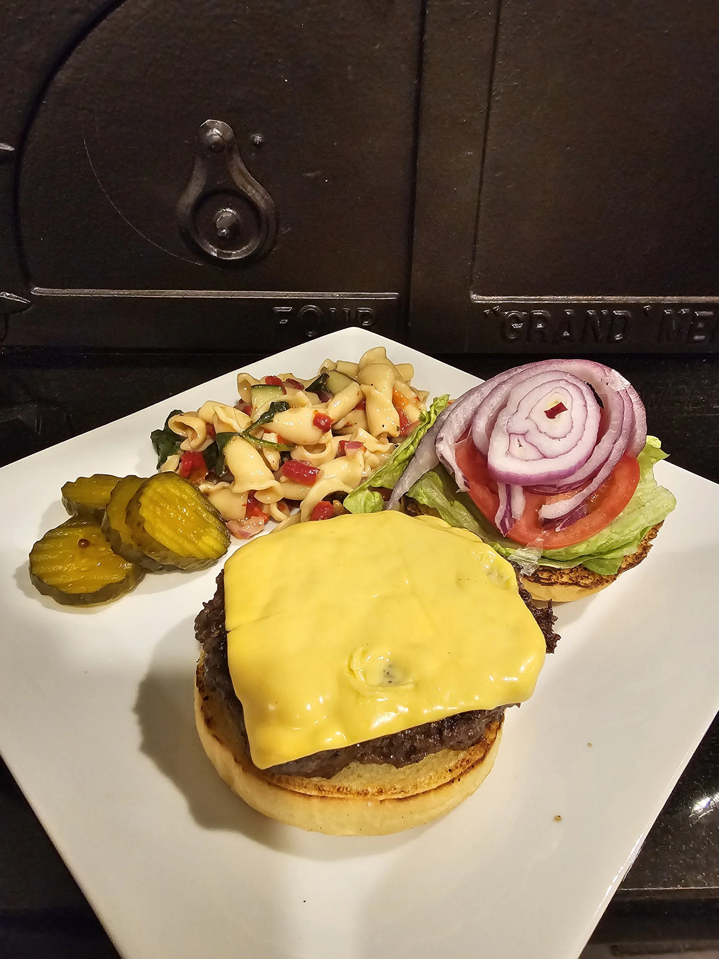 Close-up of a cheeseburger sandwich with melted yellow cheese, on a toasted bun, next to pickles, macaroni salad with red peppers, and vegetable slices, on a white plate with a black background.