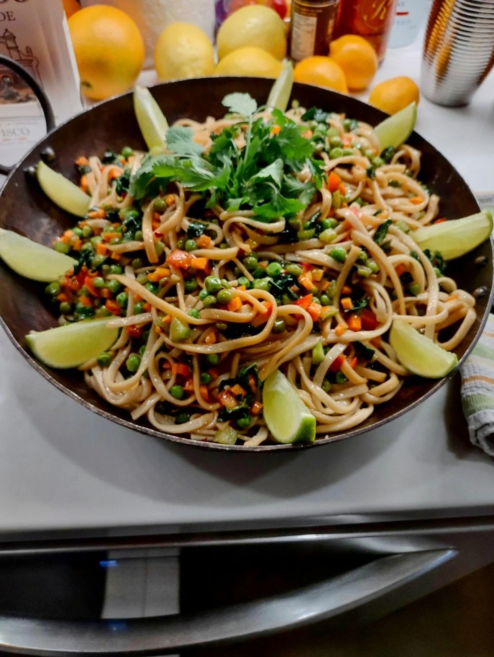 Vegetable stir-fry with noodles garnished with cilantro and lime wedges in a black skillet on a stovetop.