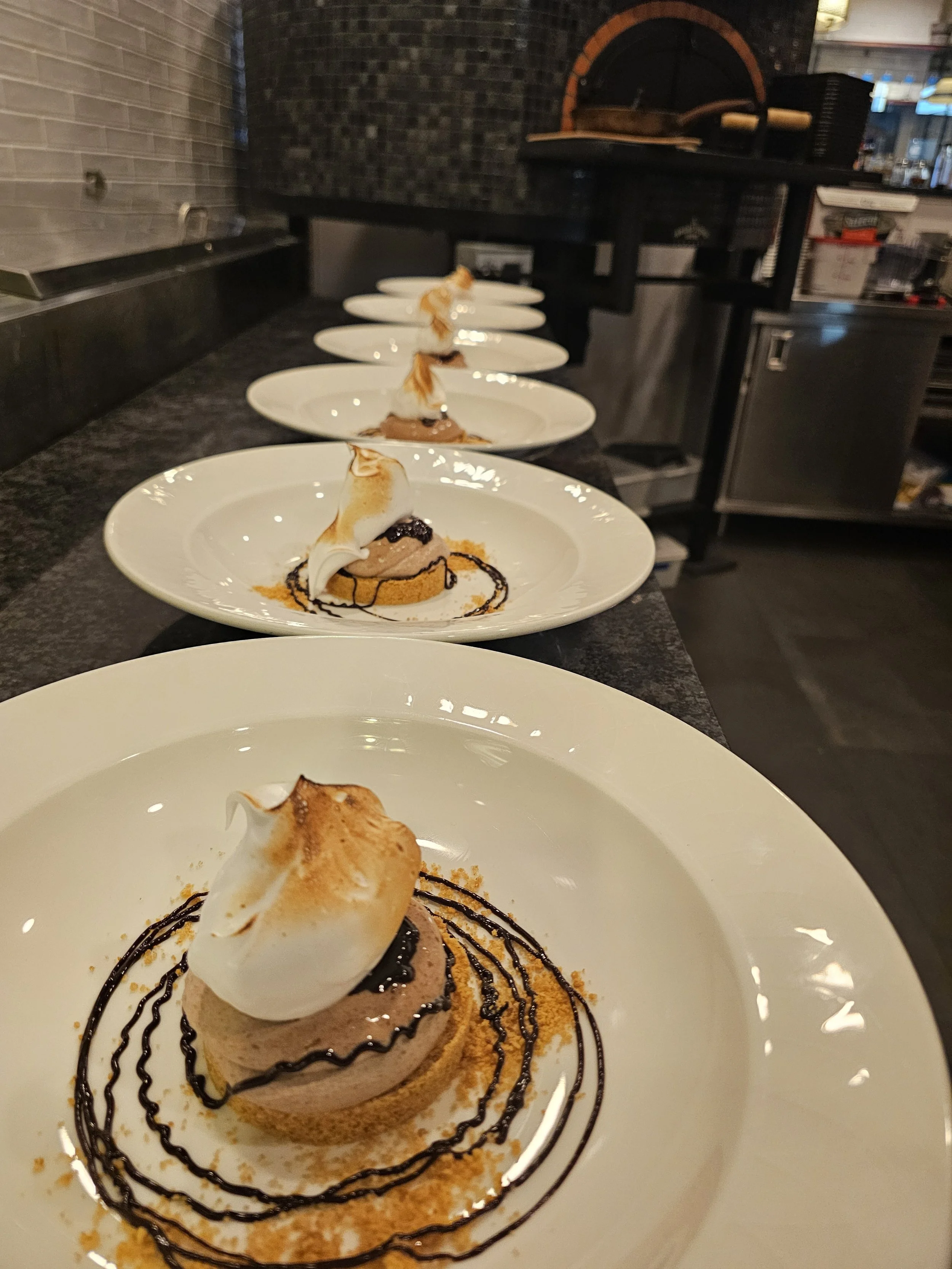 A dessert plate with a chocolate mousse topped with a toasted marshmallow, drizzled with chocolate sauce, and garnished with a sprinkle of orange-colored crumbs.