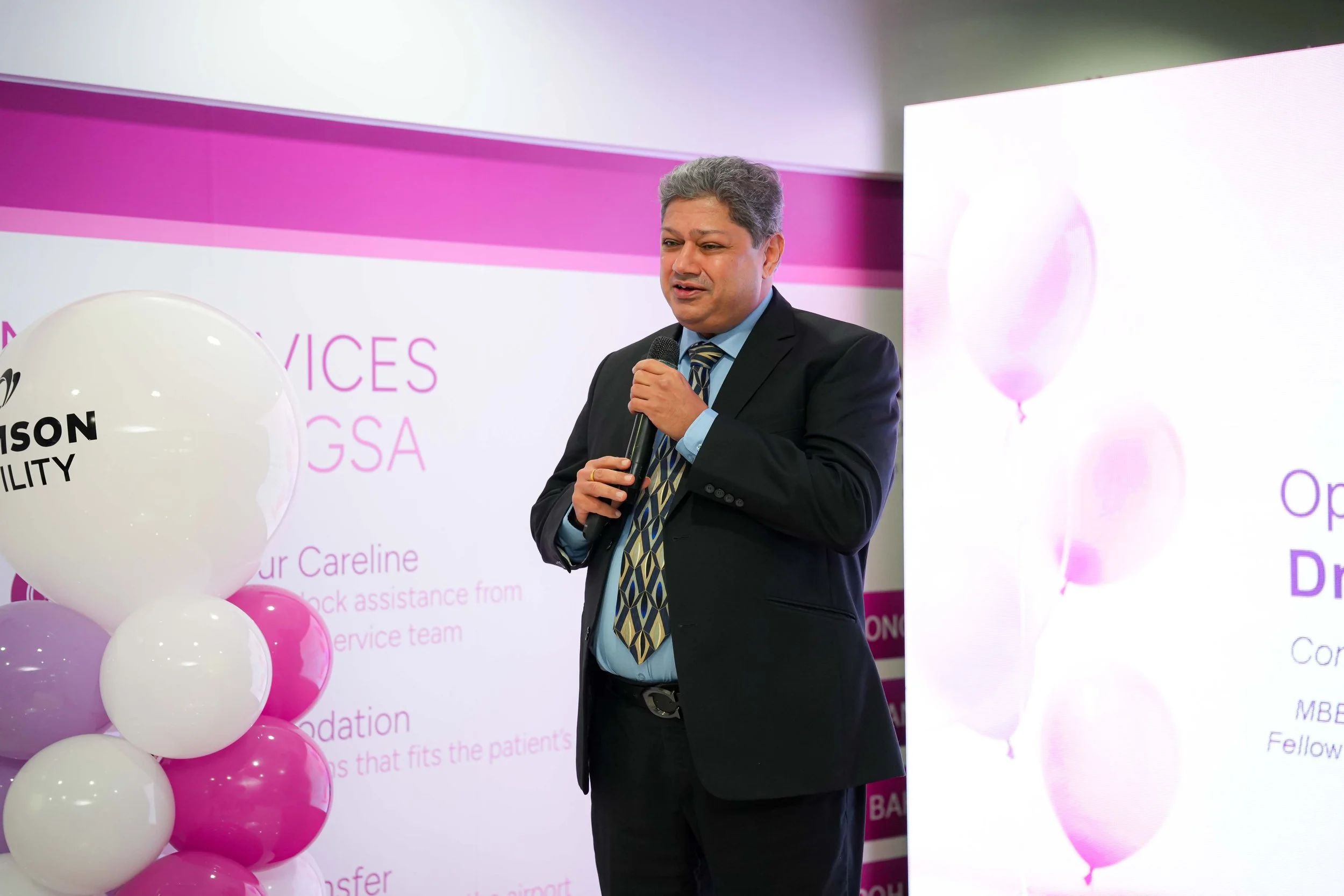 A man in a suit standing with a microphone in his hand, speaking at an event with pink and white balloons and a large pink and white presentation screen in the background.