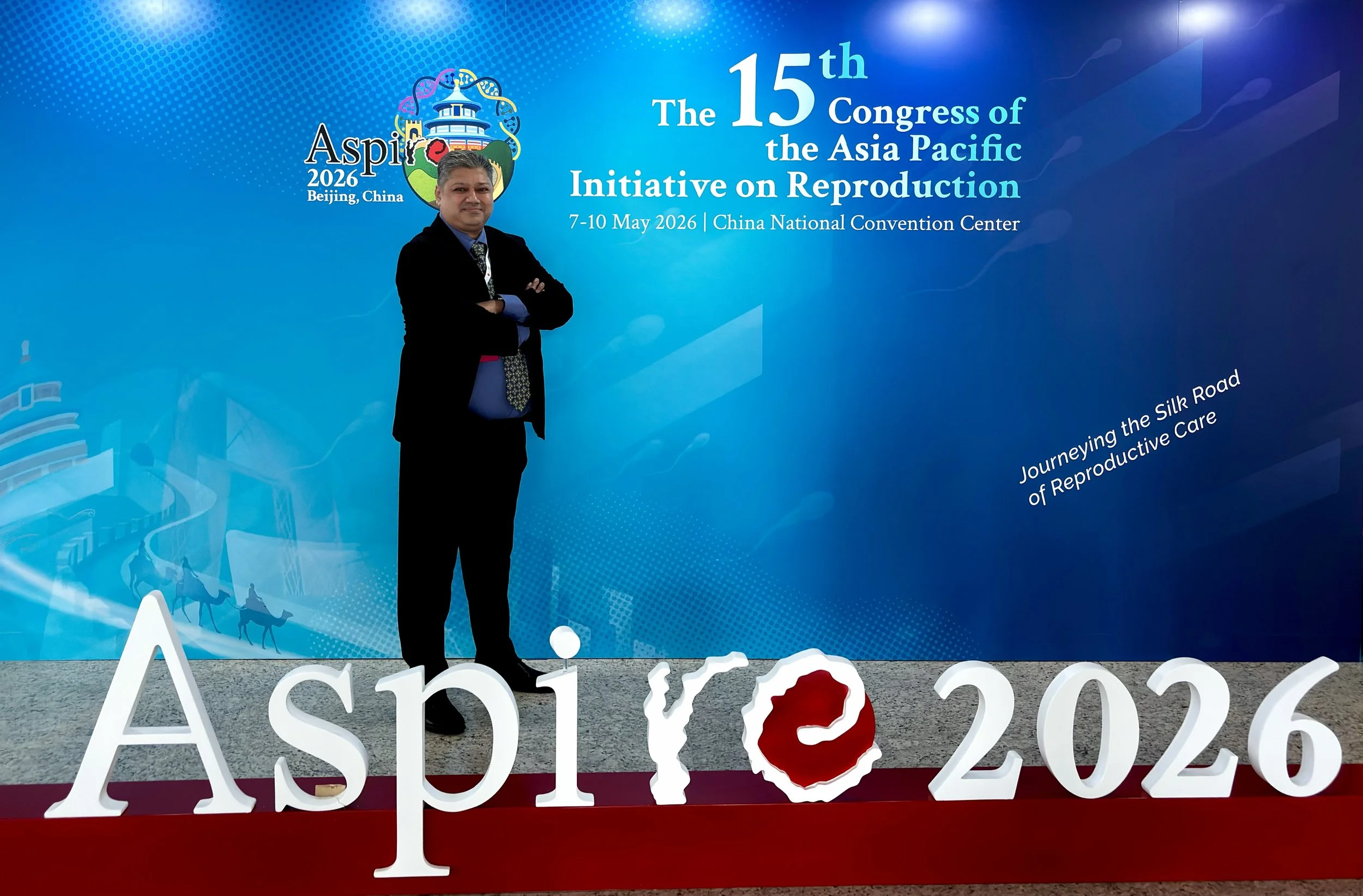 A man in a black suit stands with arms crossed in front of a blue backdrop at the 15th Congress of the Asia Pacific Initiative on Reproduction, held in Beijing, China, in May 2026. The backdrop includes a logo with a rainbow-colored DNA helix, a pagoda, and a landscape. Decorative white lettering spelling 'Asia,' and partial letters of other words are visible on the floor.
