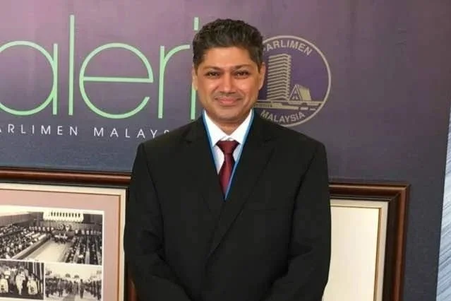 Man in a black suit with a white shirt and red tie, standing in front of a purple background with the words partially visible and a logo.