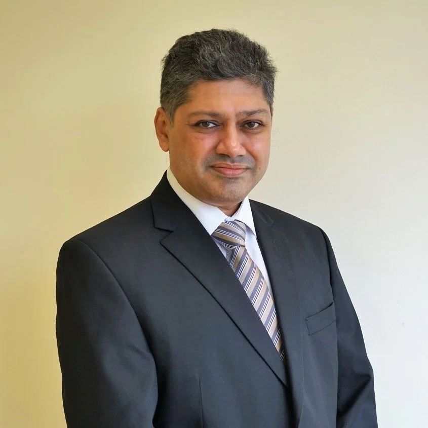 Professional man in a black suit with a striped tie posing against a beige background.
