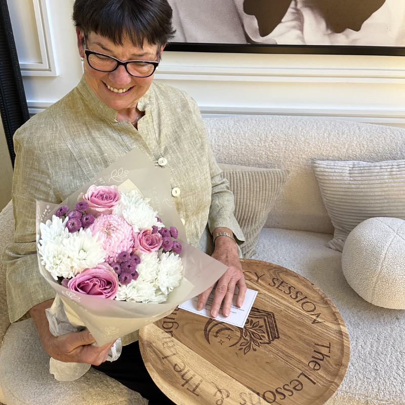 A woman with short dark hair and glasses is smiling and holding a bouquet of pink, white, and purple flowers while sitting on a beige couch in a room with decorative pillows.