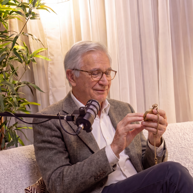 Older man with glasses in a gray blazer looking at a pocket watch, sitting on a sofa with cream-colored curtains and a potted plant in the background.